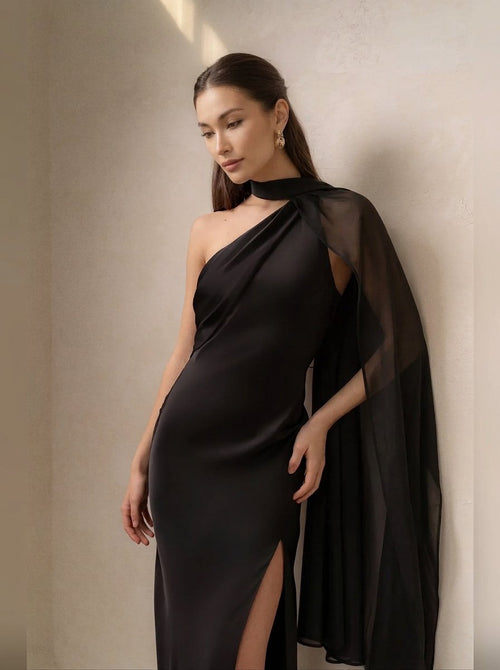 A woman wearing a black one-shoulder satin gown with a sheer draped cape and a thigh-high slit, posing against a textured beige wall.