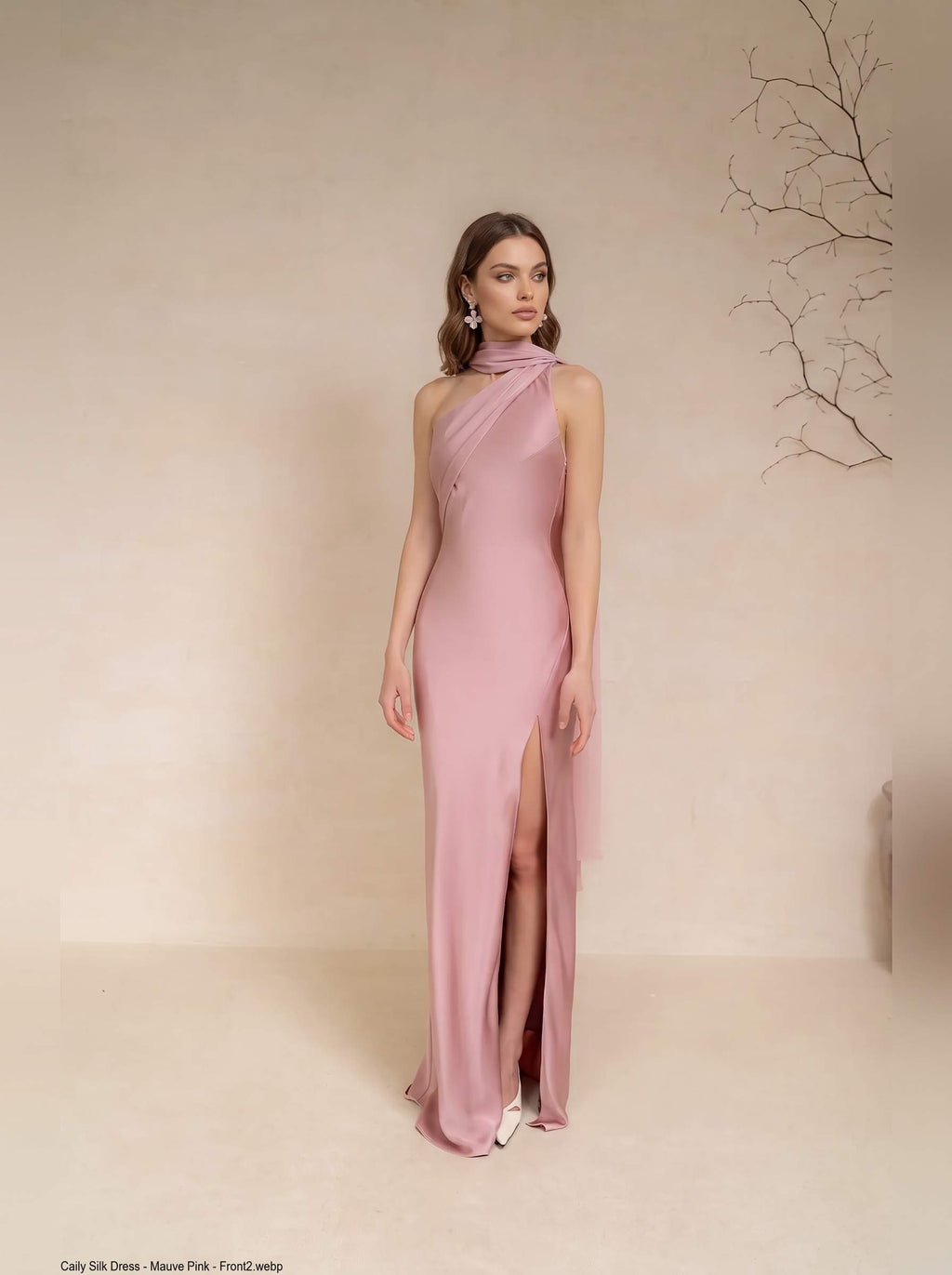 A woman models a floor-length dusty pink satin gown with a high slit and draped halter-style neckline against a warm beige studio backdrop.