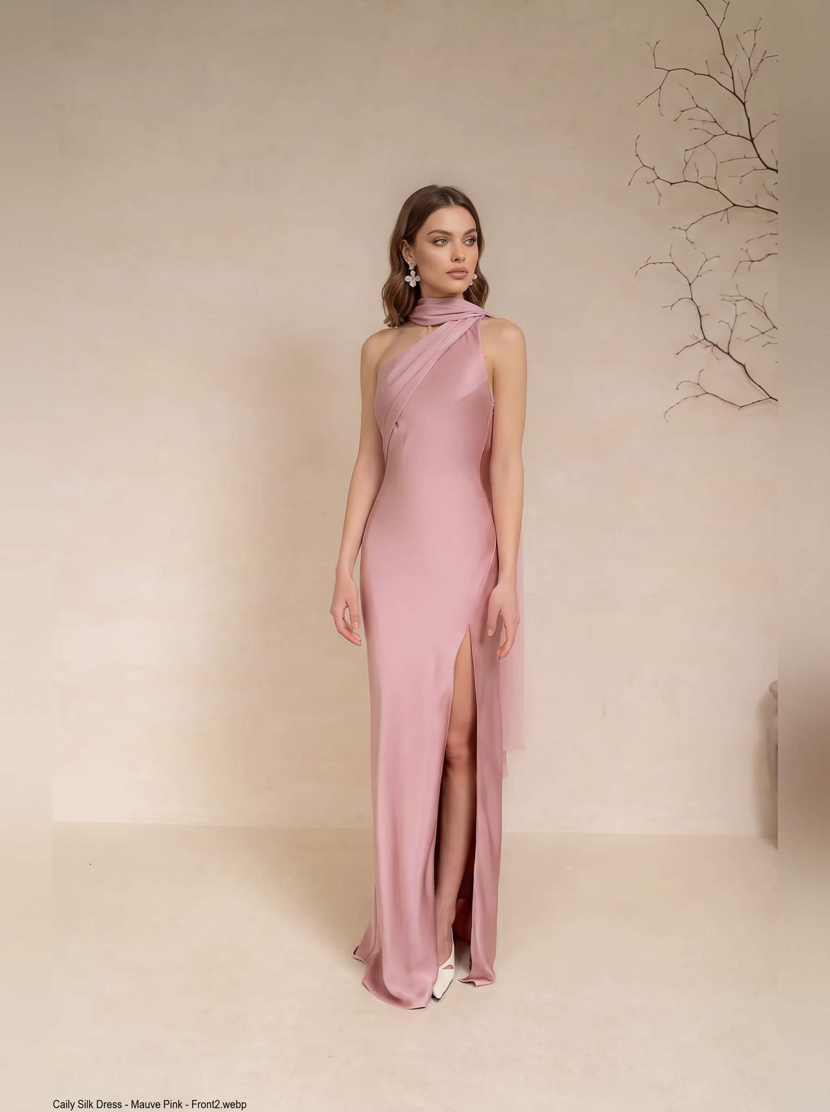 A woman models a floor-length dusty pink satin gown with a high slit and draped halter-style neckline against a warm beige studio backdrop.