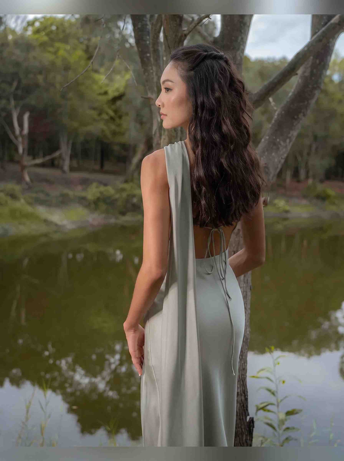 A woman stands outdoors by a lake wearing a sleeveless, floor-length sage green dress with an open back and tie detail. Trees and reflective water are visible in the background.