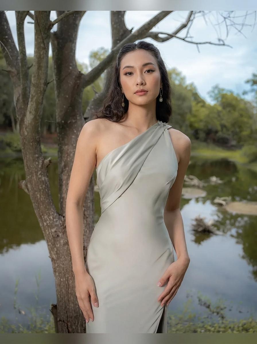 A woman wearing a draped, one-shoulder sage green dress stands outdoors next to a tree by a calm lake.