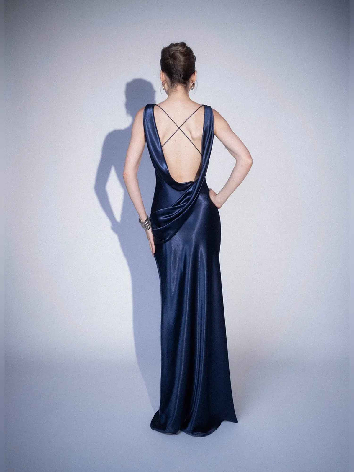 A woman stands with her back to the camera wearing a floor-length navy satin gown with a deep open back and crisscross strap detail, photographed against a light gray studio background.