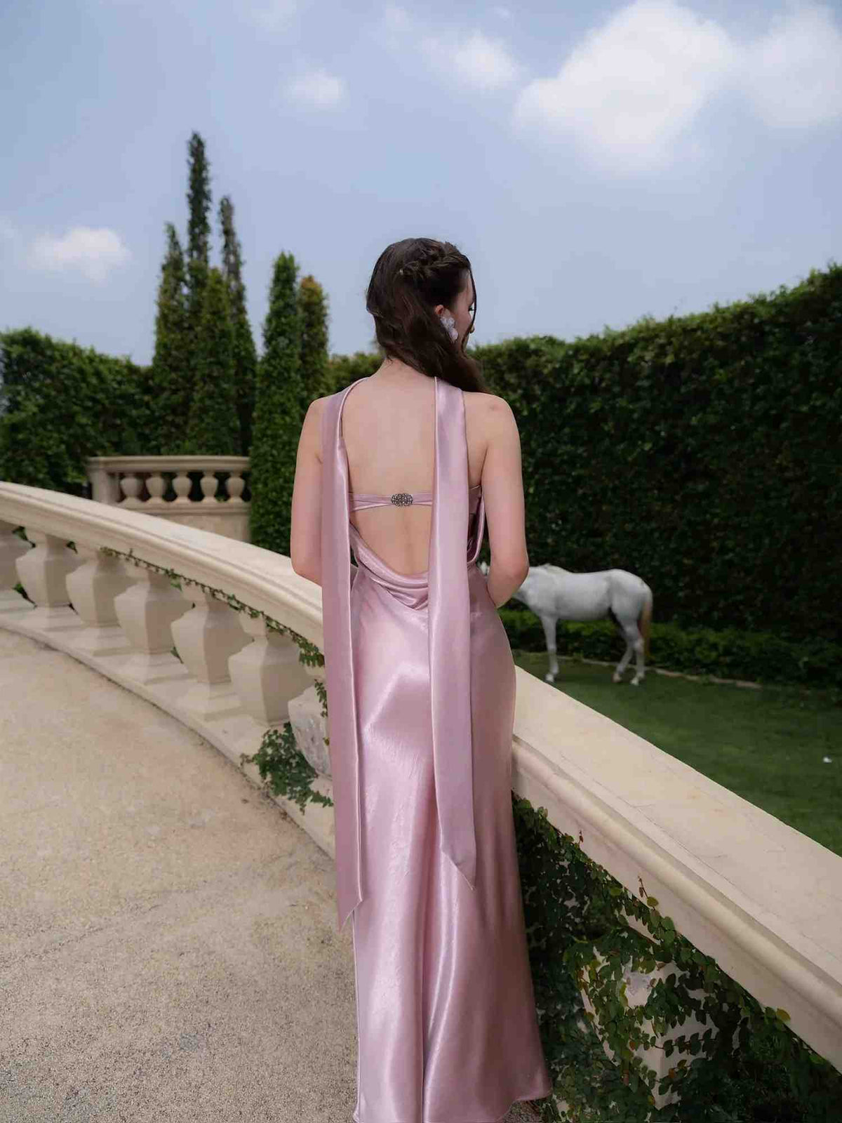 A woman stands on a stone terrace outdoors, viewed from behind, wearing a floor-length satin gown in a dusty pink color. The dress features a low open back with a long draped tie and delicate shoulder straps.