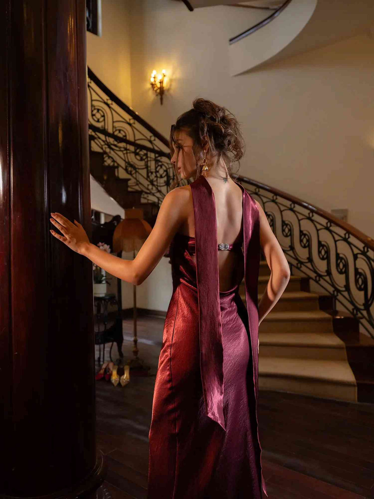 A woman in a shimmering deep burgundy satin evening gown with a draped back detail, posing indoors next to a dark wooden pillar in front of a staircase.