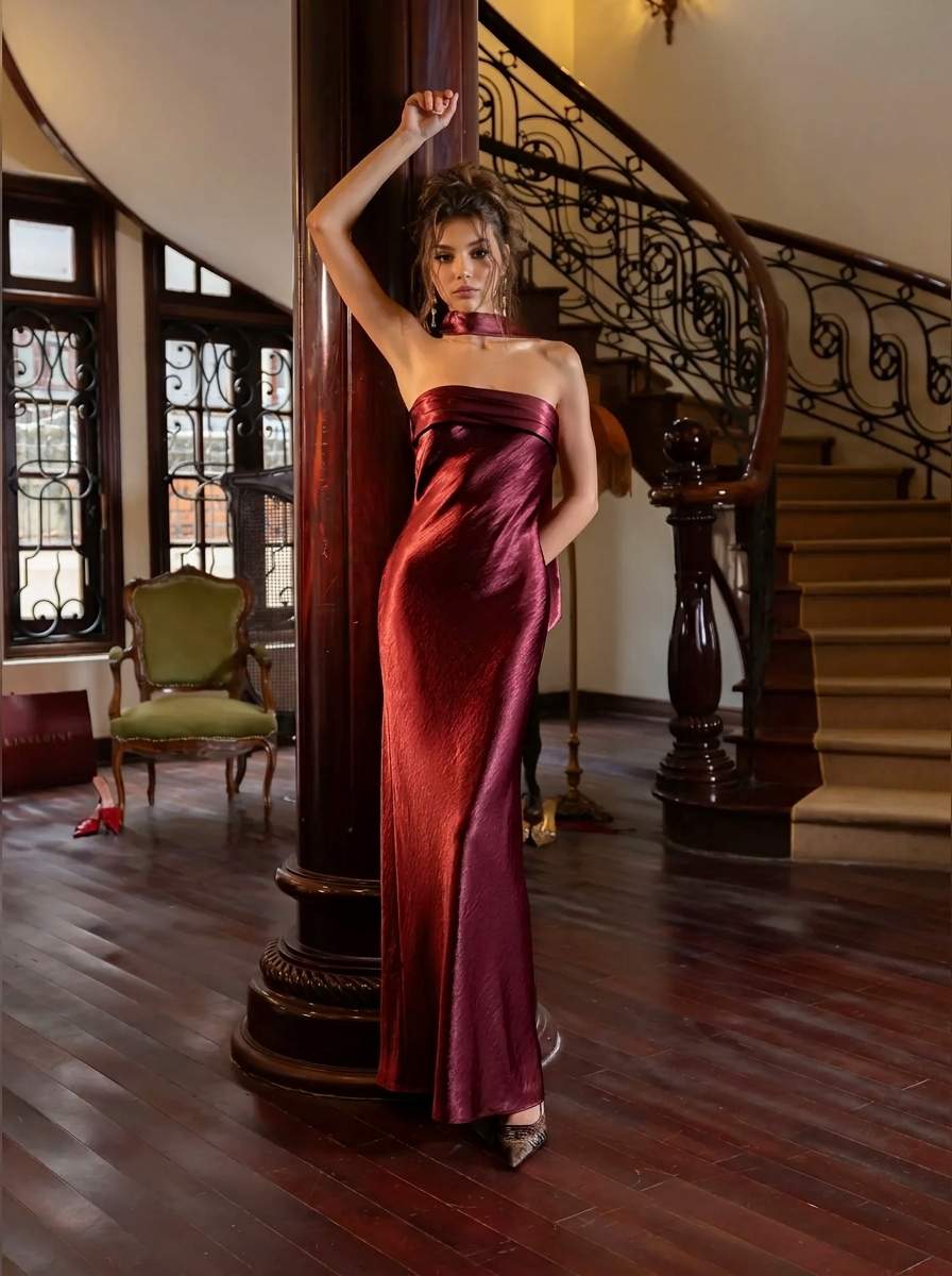 A woman models a strapless metallic burgundy maxi dress with a high neck and thigh-high slit, posing in a grand interior with a wooden staircase and dark wood columns.