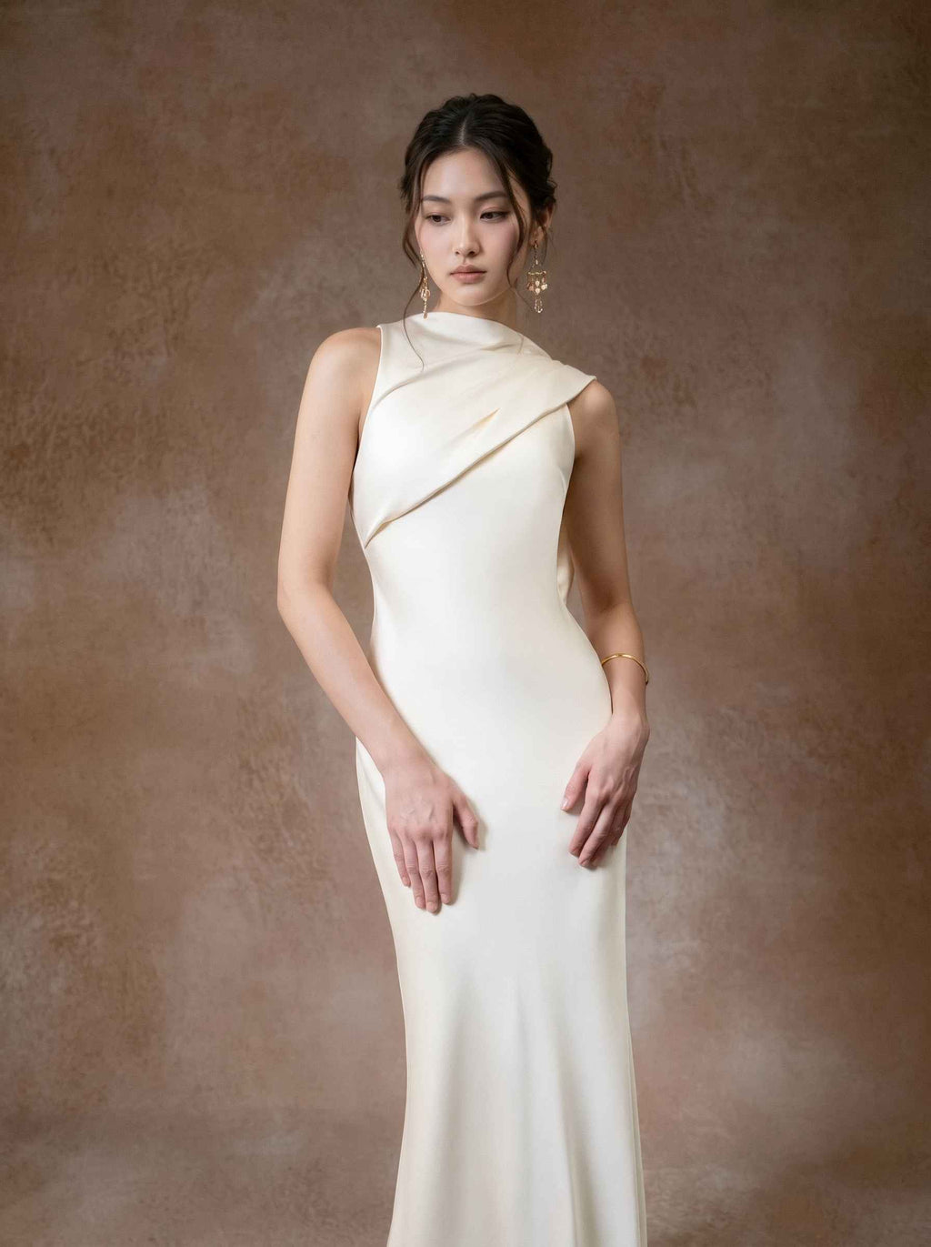 A woman modeling a sleeveless, off-white evening gown with a draped, asymmetrical neckline and a fitted silhouette, posed against a textured brown studio backdrop.