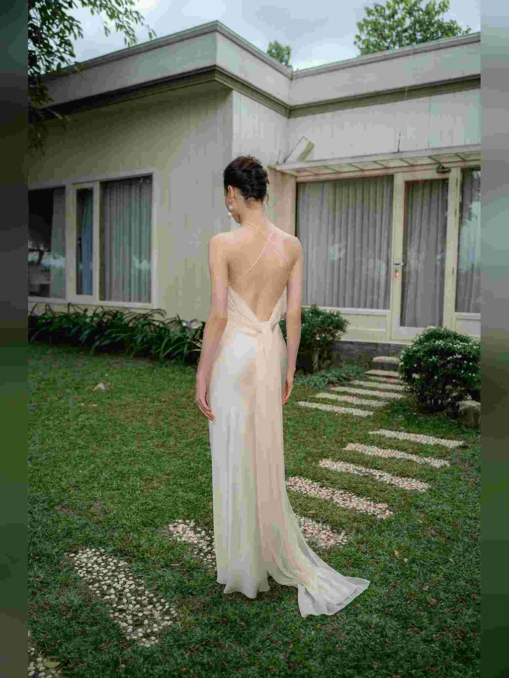A woman stands outdoors in a garden courtyard wearing a flowing backless gown, photographed from behind. The dress is a pale blush or champagne tone with a draped open back and a long train.