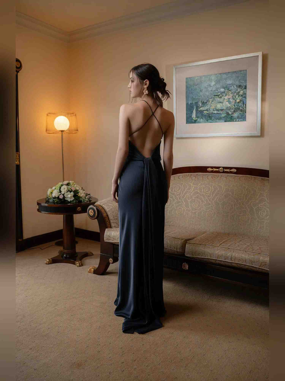 A woman stands in a warmly lit room wearing a floor-length navy evening gown with a low open back and thin shoulder straps, photographed from behind.