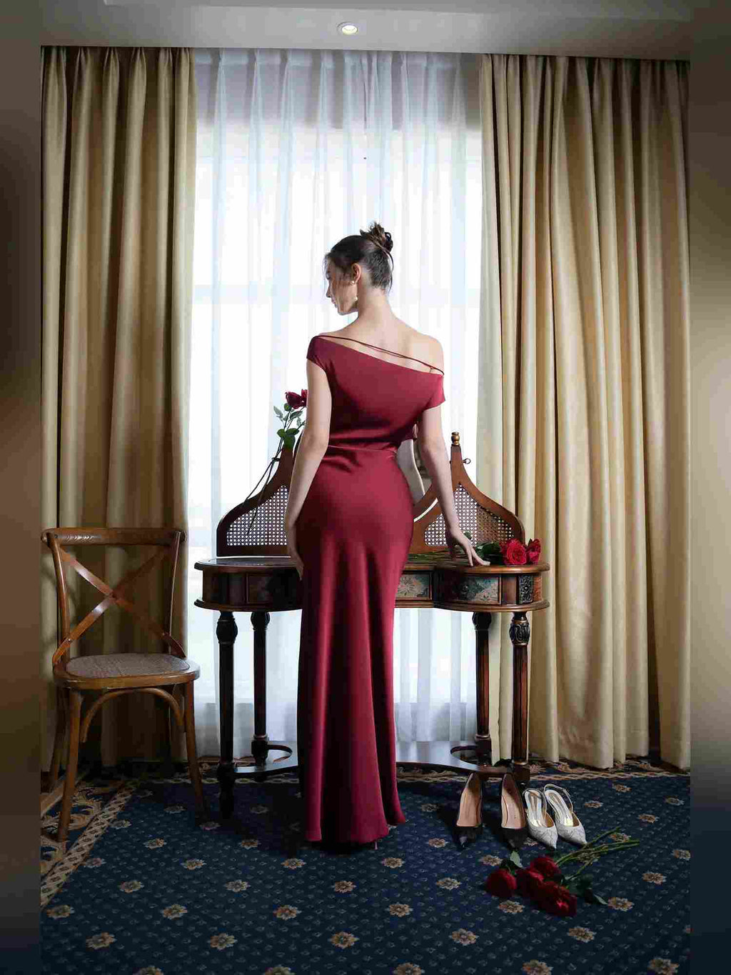 A woman stands with her back to the camera in a luxurious room, wearing a floor-length, off-the-shoulder burgundy evening gown. She is positioned in front of a window with sheer white curtains and heavy gold drapes, with a wooden console table, chairs, and red roses in the background.
