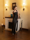 A woman models a floor-length black satin gown with a one-shoulder strap design in a warm, vintage-style room with a fireplace and wall sconces.