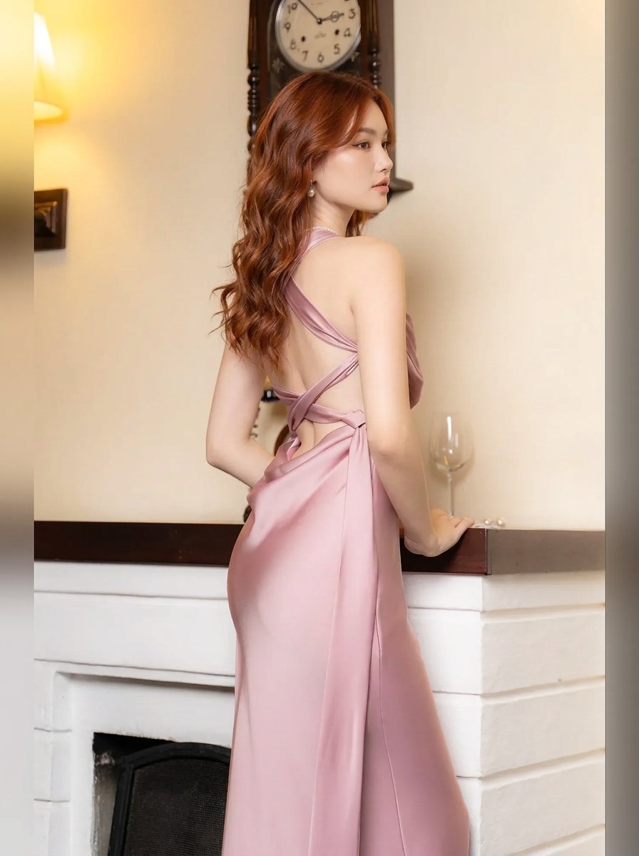 A woman with long wavy red hair models a dusty pink satin halter-neck dress with a backless crossover design, standing in front of a fireplace.