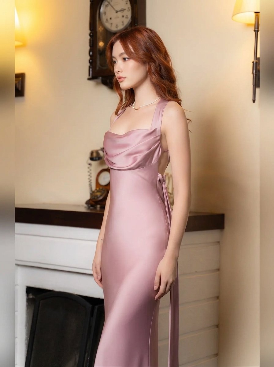 A woman wearing a satin-like dusty pink evening gown with a draped bodice and side tie detail, standing in a warmly lit indoor room with a fireplace and wall sconces.