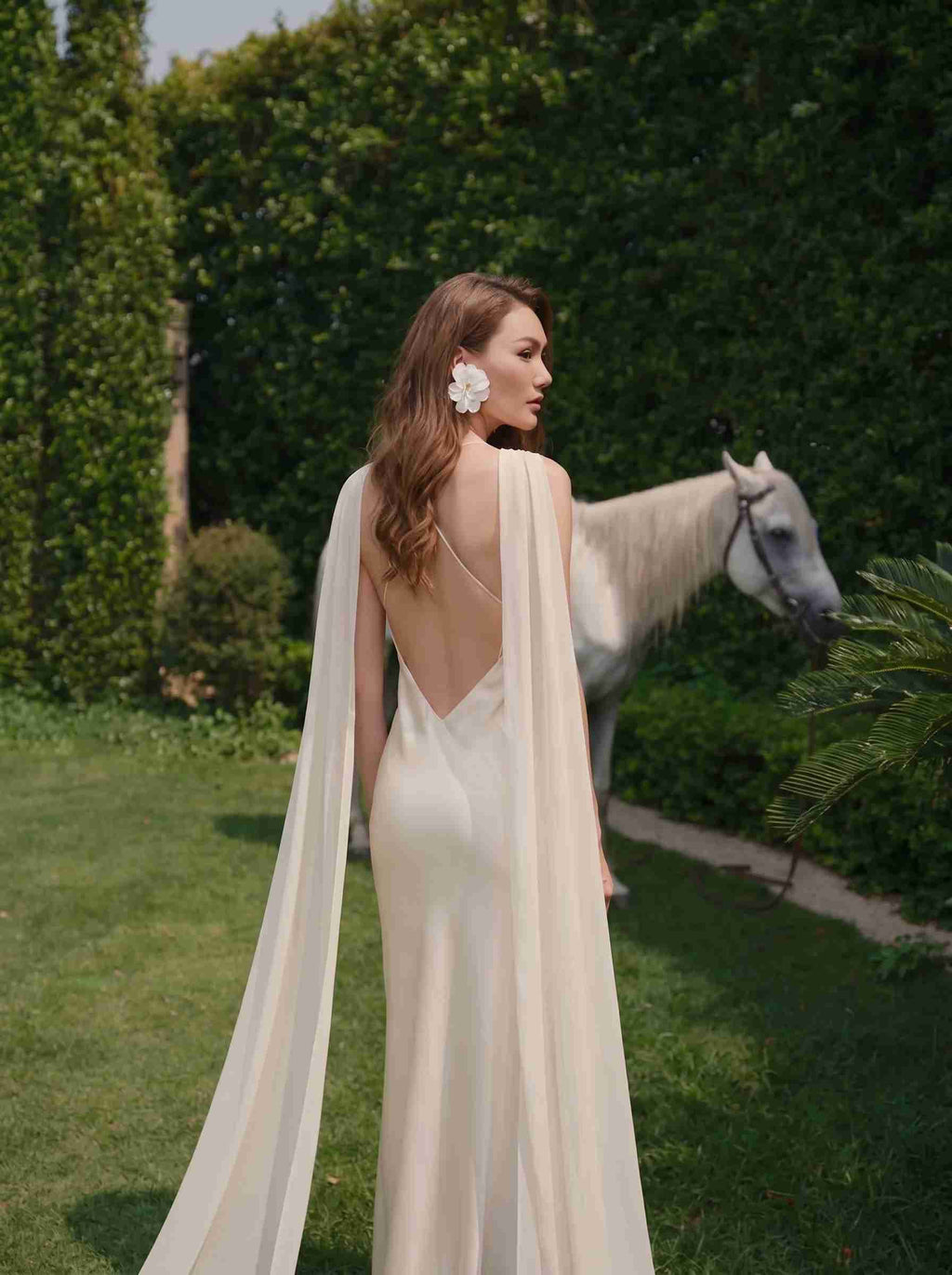 A woman stands outdoors in a lush garden wearing a floor-length cream gown with a draped cape-style back and open back. A white horse is visible in the background among green hedges and grass.