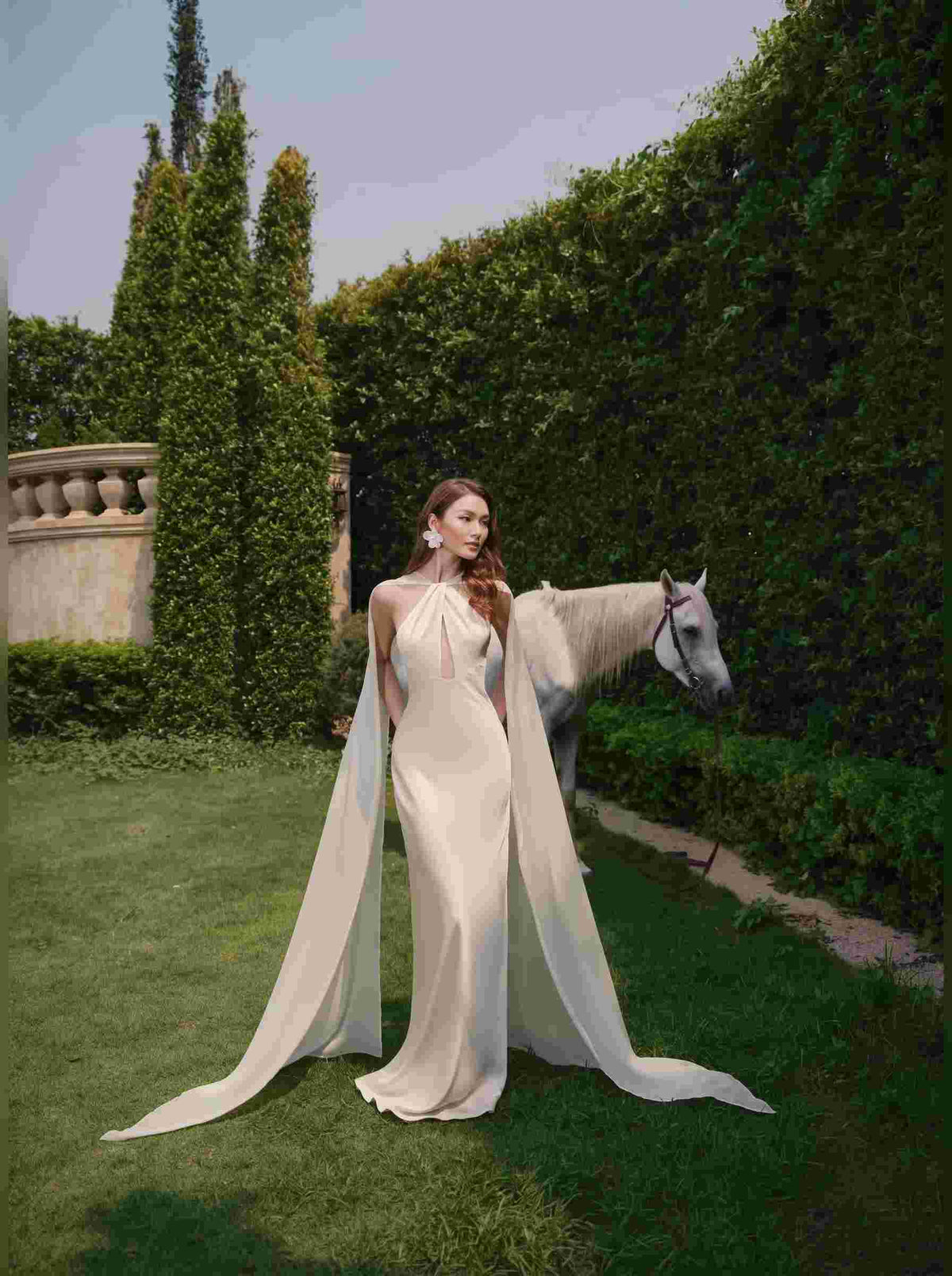 A woman stands in a lush garden wearing a floor-length cream gown with a dramatic flowing sheer train, while a white horse stands beside her against a backdrop of green hedges and grass.