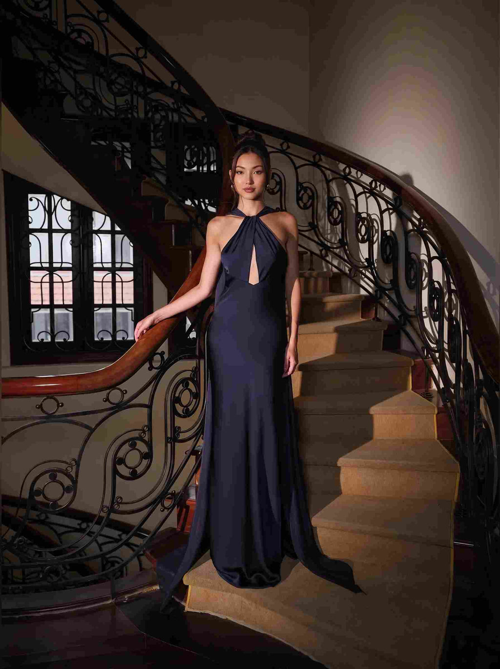 A woman wearing a floor-length navy evening gown with a halter neckline and keyhole cutout stands on the steps of an ornate indoor staircase with a curved wooden handrail and black metal railing.