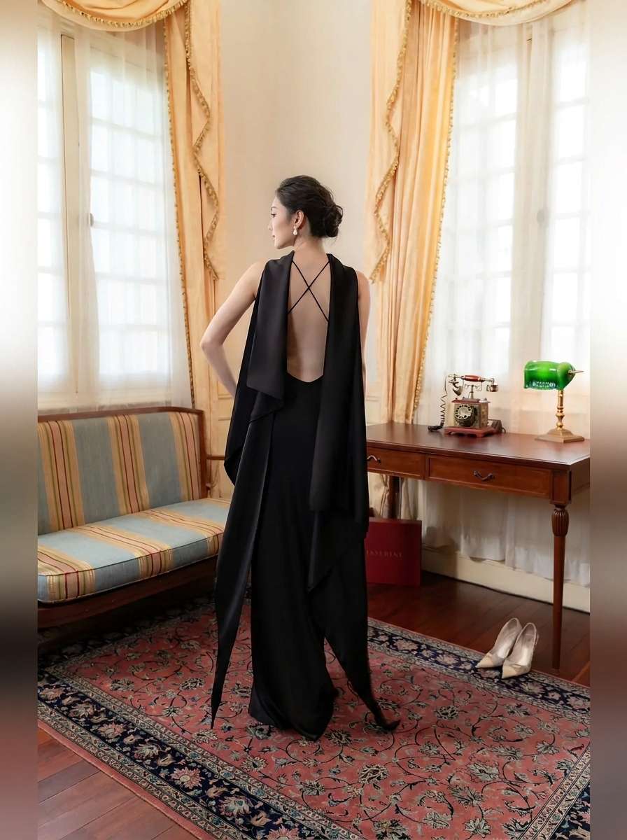A woman stands in a vintage room wearing a long black dress with a dramatic open back and draped overlay. The room features cream curtains, a striped bench, a wooden desk, and a patterned rug.