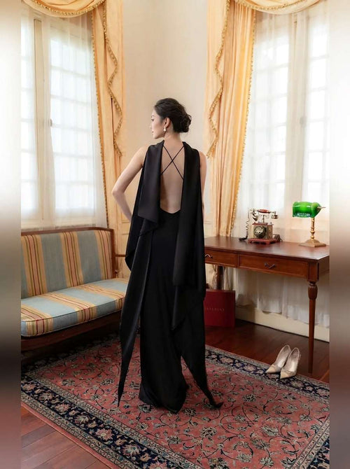 A woman stands in a vintage room wearing a long black dress with a dramatic open back and draped overlay. The room features cream curtains, a striped bench, a wooden desk, and a patterned rug.