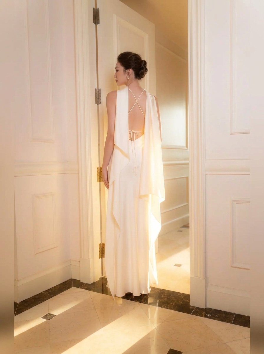 A woman stands in a doorway wearing a flowing white sleeveless gown with a draped cape-like overlay and an open back, photographed in a bright indoor hallway with cream walls and tiled flooring.