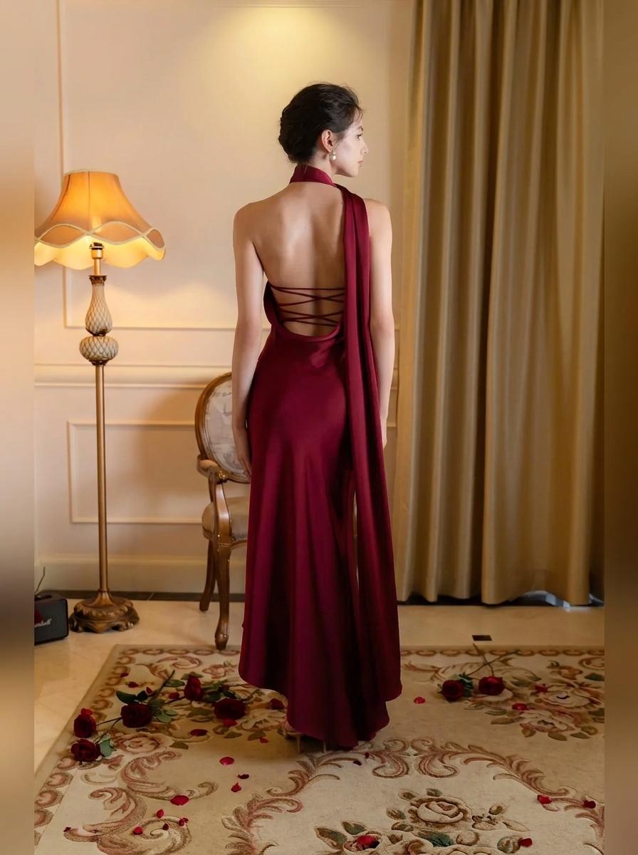 A woman stands with her back to the camera wearing a floor-length burgundy evening gown with a draped one-shoulder back design and crisscross lace-up detailing. The setting is an elegant room with a gold floor lamp, patterned curtains, and a floral rug.