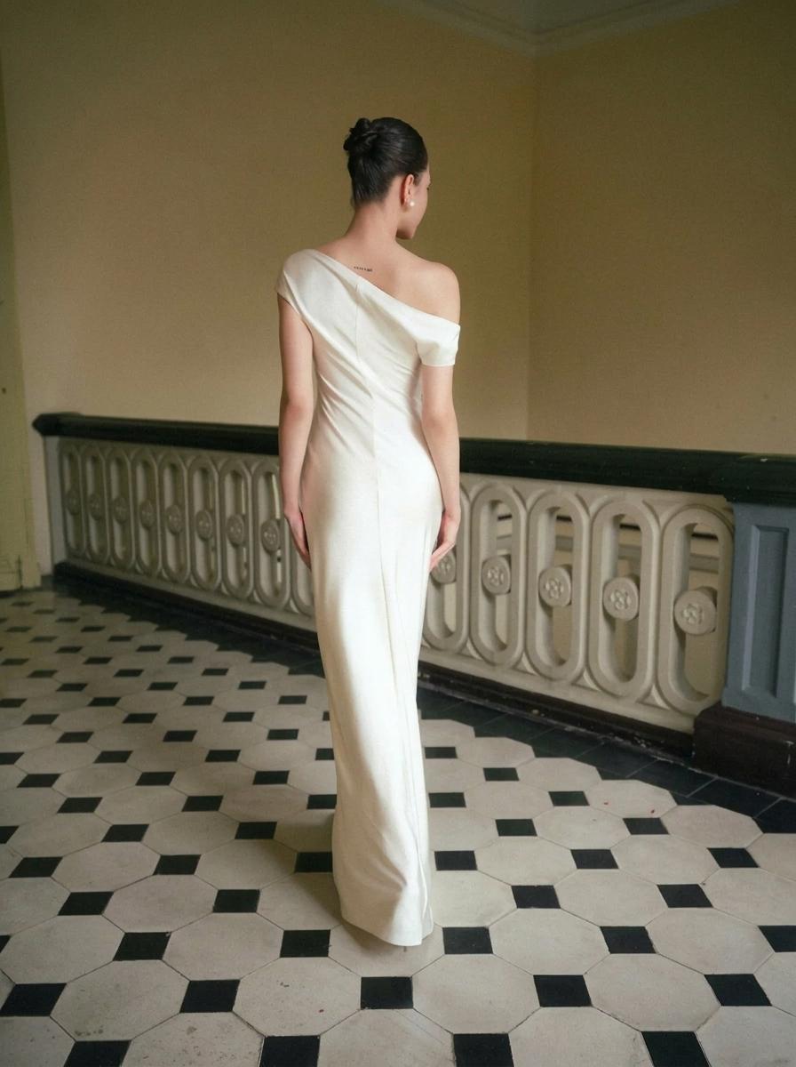 A woman stands with her back to the camera wearing a floor-length off-the-shoulder ivory gown in an indoor hallway with a black-and-white tiled floor and a decorative balustrade.