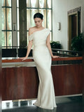 A woman wearing a floor-length off-the-shoulder ivory gown with a fitted silhouette and subtle sheen, leaning against a wooden railing in a modern indoor setting.