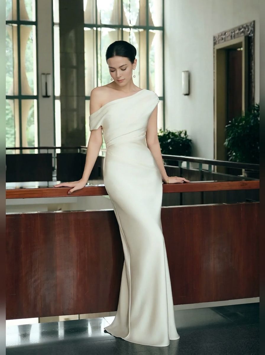 A woman wearing a floor-length off-the-shoulder ivory gown with a fitted silhouette and subtle sheen, leaning against a wooden railing in a modern indoor setting.