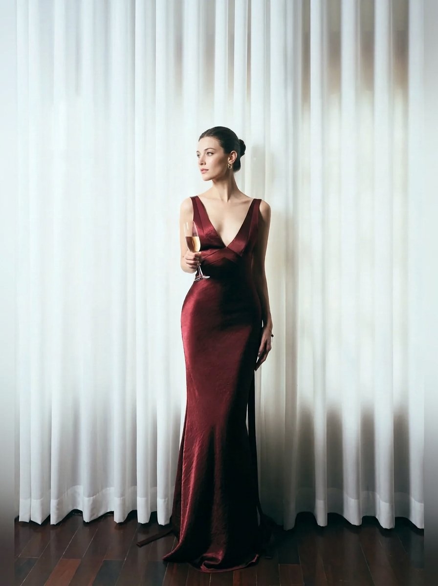 A woman stands in front of white draped curtains wearing a floor-length deep burgundy satin gown and holding a glass of white wine.