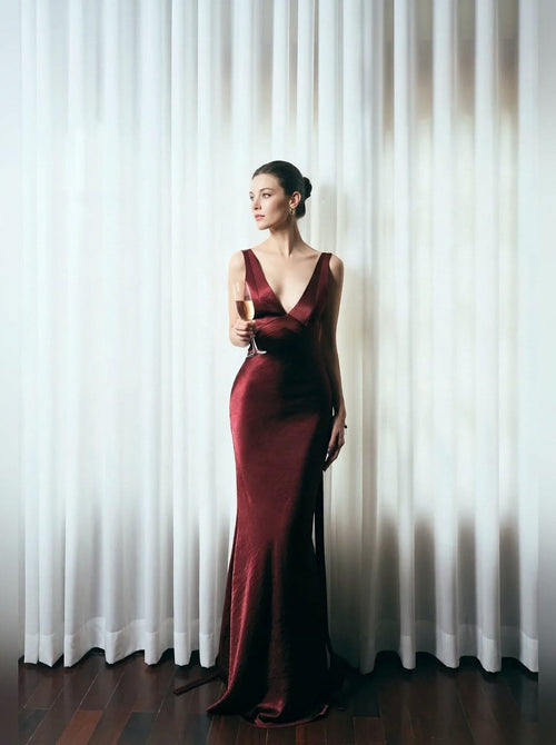 A woman stands in front of white draped curtains wearing a floor-length deep burgundy satin gown and holding a glass of white wine.